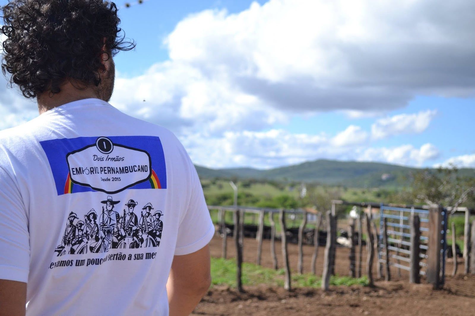 Igor de camisa do Empório Pernambucano diante da paisagem do sertão, com cerca e curral